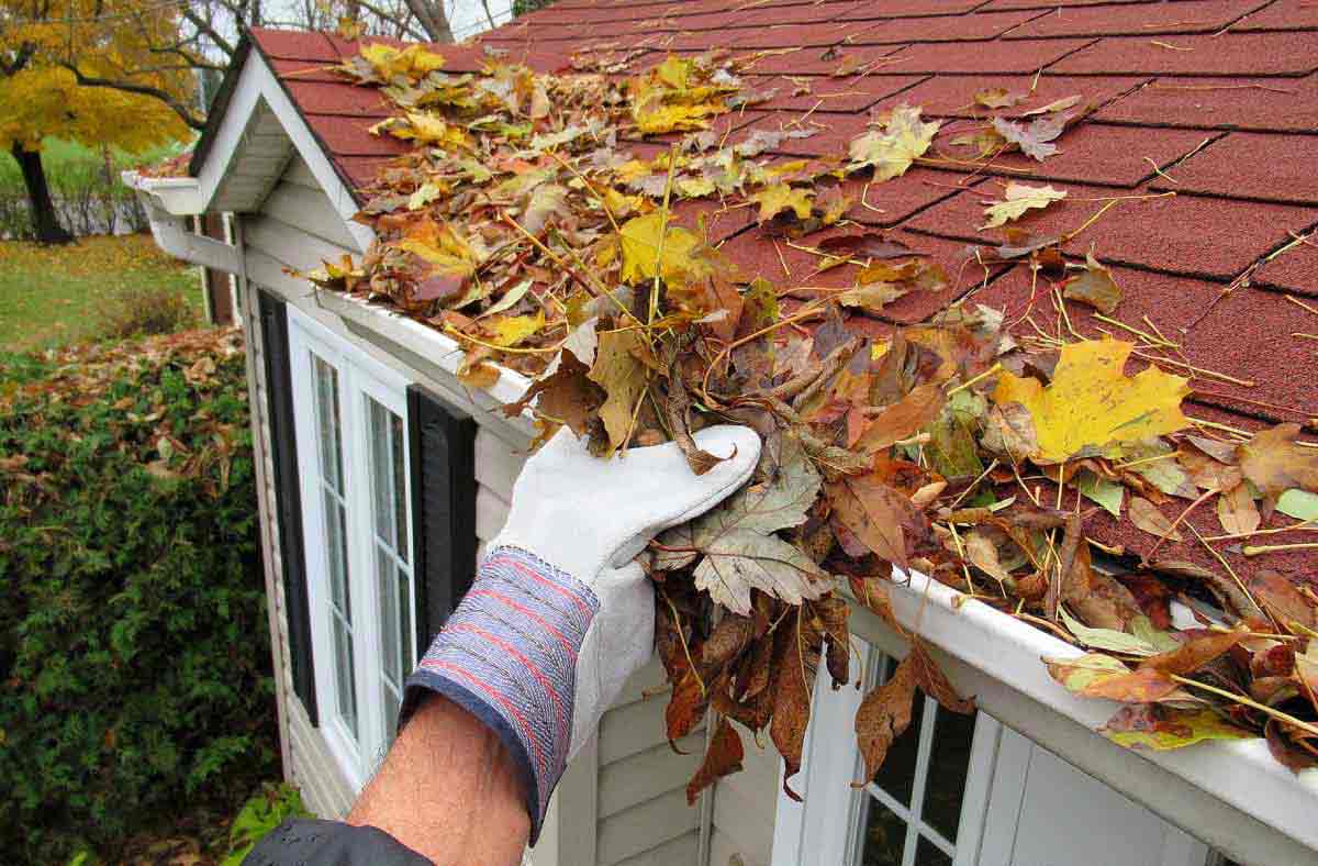 A gloved hand reaches up to clear leaves out of a roof gutter